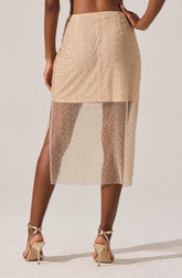 The image shows a woman wearing a beige skirt with a high waist and a fitted silhouette. The skirt has a dual-layer design: the top layer is solid and opaque, ending above the knee, while the bottom layer is made of sheer, see-through fabric extending to mid-calf length. The sheer portion has a dotted pattern, creating a semi-transparent effect. She is also wearing beige high-heeled sandals with ankle straps.